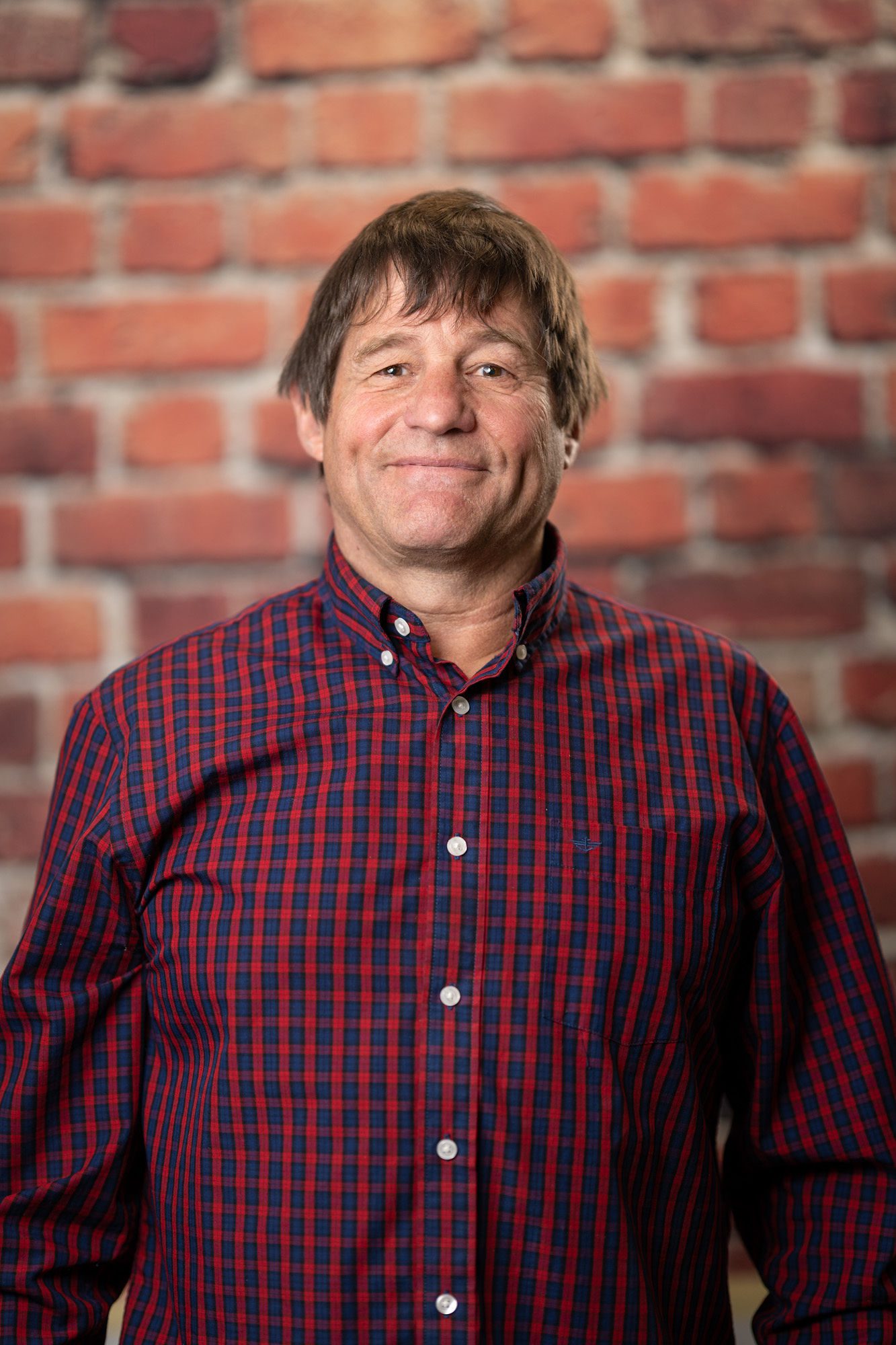 A middle-aged person with medium-length brown hair wearing a red and navy plaid button-up shirt stands in front of a brick wall.