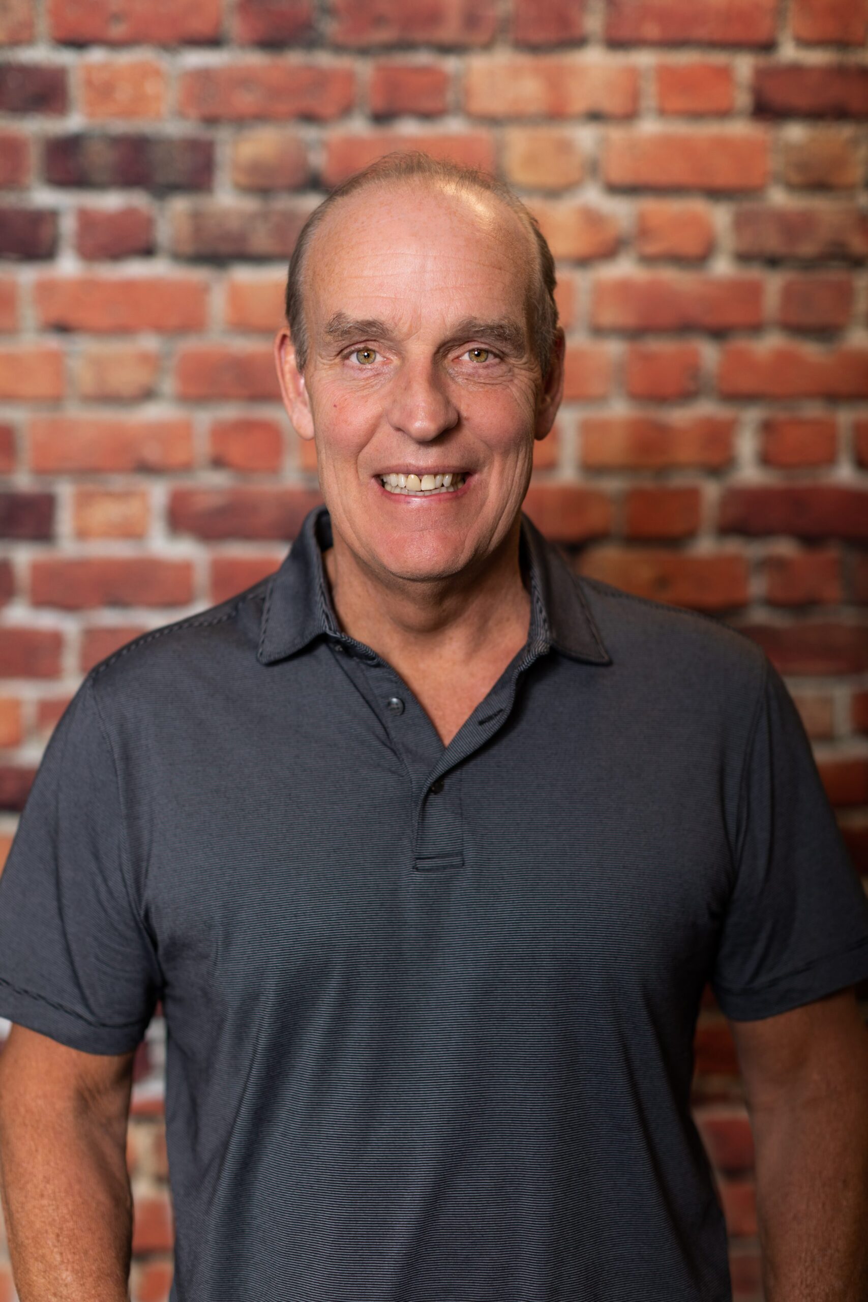 A man with short hair and a trimmed beard wearing a light blue, short-sleeved button-up shirt stands against a background of red brick.